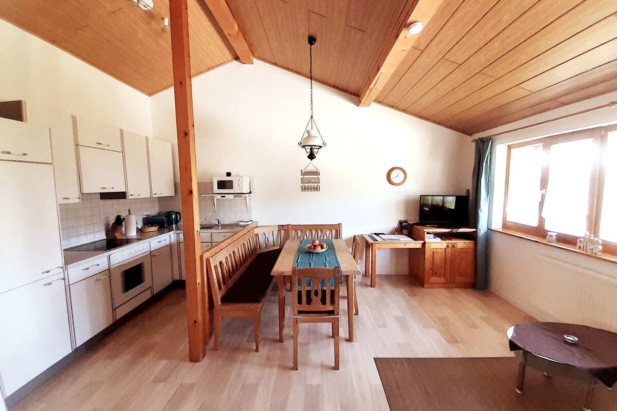 A spacious kitchen and dining area is provided with wooden beams and a high ceiling. A large table with benches sits in the center, accompanied by a stove, oven, and other essential appliances. Natural light brightens the room through large windows.