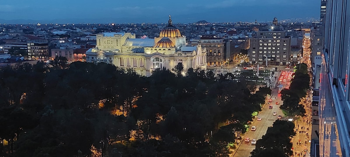 An evening view showcases the illuminated Palacio de Bellas Artes, with its distinctive dome gleaming against the city skyline. The bustling streets below are filled with traffic, while trees and green spaces provide a contrast to the urban environment.