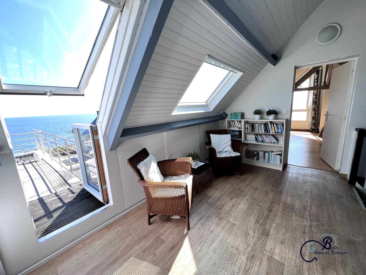 A serene corner in an upper-level room features two wicker chairs positioned near large, angled windows with ocean views. A light wooden floor complements a nearby bookshelf lined with books, while natural light fills the airy space.
