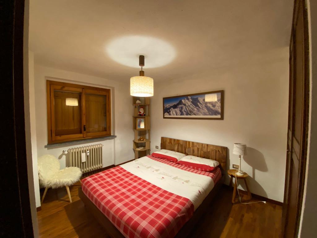 A cozy bedroom features a spacious bed with red and white checked bedding. A framed mountain picture adorns the wall above the bed. Natural light enters through a window with wooden shutters, while a soft chair and modern lamp provide additional comfort.
