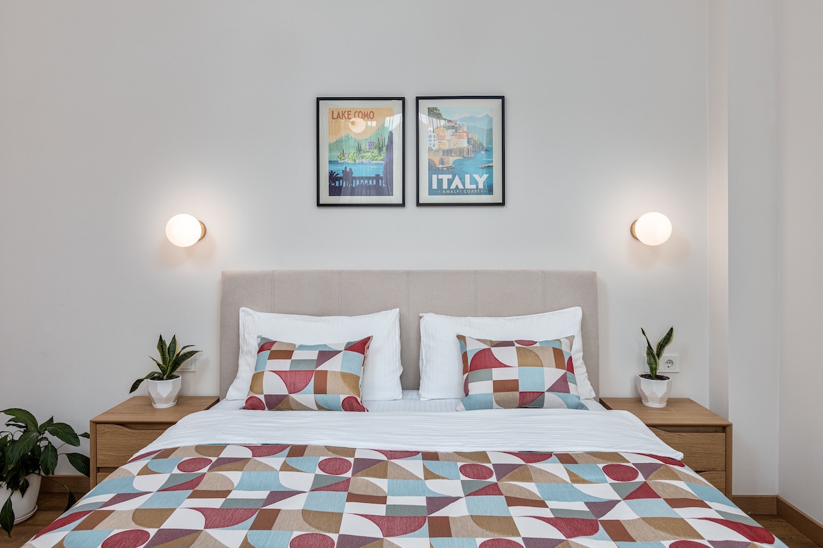 A spacious bed is centered against a light-colored wall, featuring decorative pillows and a patterned bedspread. Above the bed, two framed prints add a touch of color. Small potted plants are placed on nightstands, enhancing the serene atmosphere of the room.