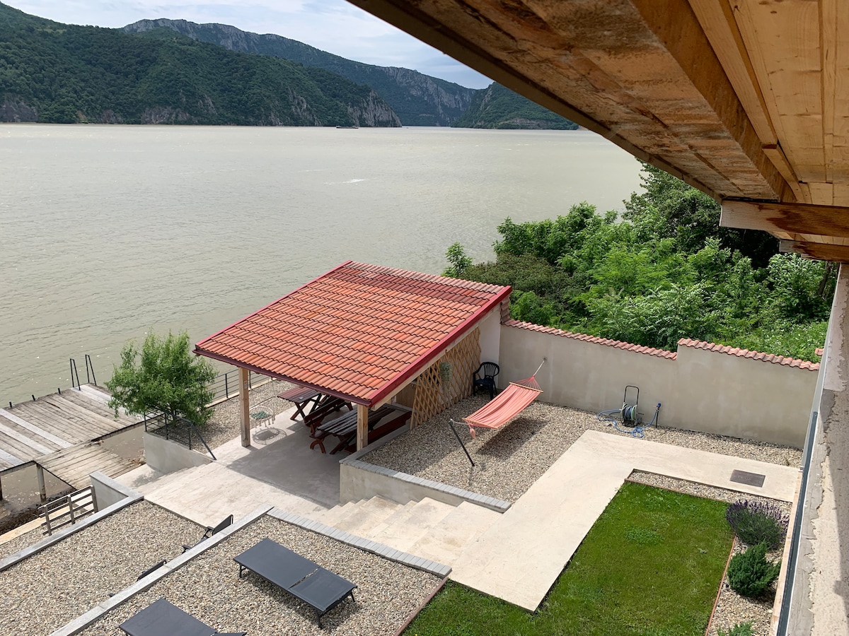 A serene outdoor space is visible with a covered pavilion featuring a red-tiled roof. It includes a dining area and a hammock. Below, a well-maintained garden area complements the natural surroundings, with a clear view of the river and distant mountains.