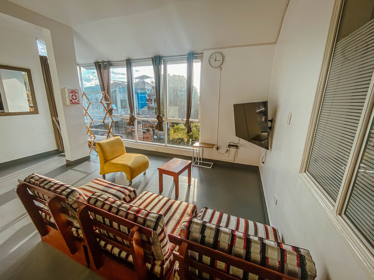 A living area features large windows allowing natural light to fill the space. A striped sofa and a yellow chair provide seating, accompanied by a small table. A wall clock and a television are visible, contributing to the functional layout.