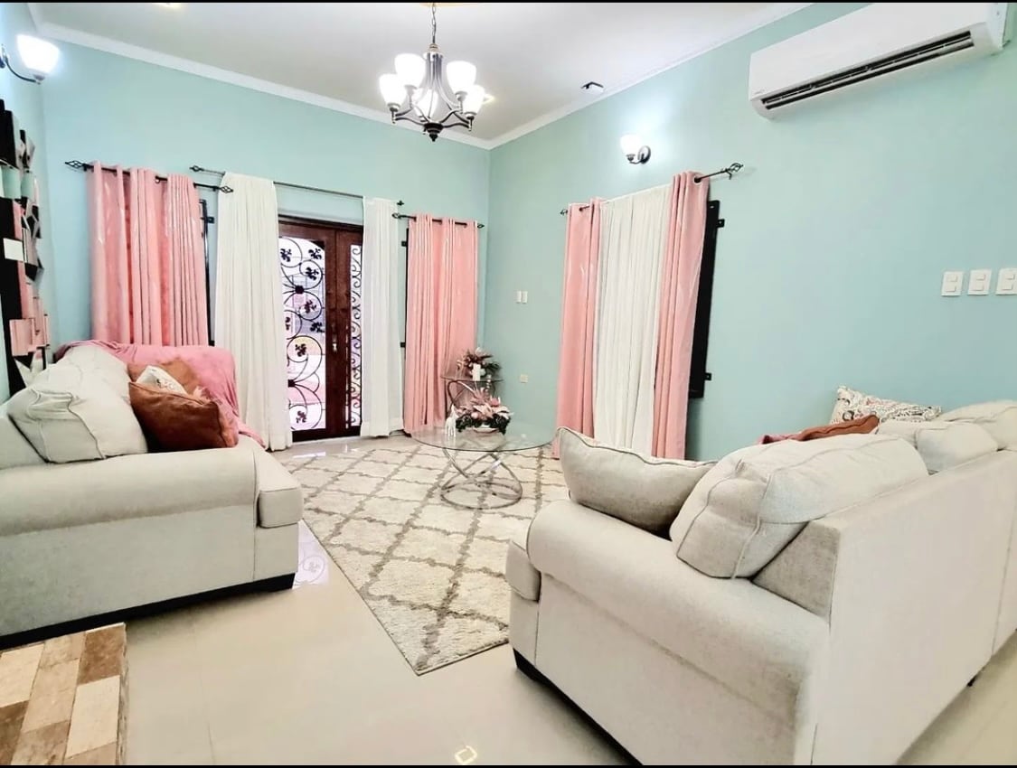 A cozy living area is presented with soft, neutral-toned sofas placed around a decorative coffee table. Light pink and white curtains frame the large entrance, allowing natural light to brighten the space. A patterned rug adds texture to the well-lit room.
