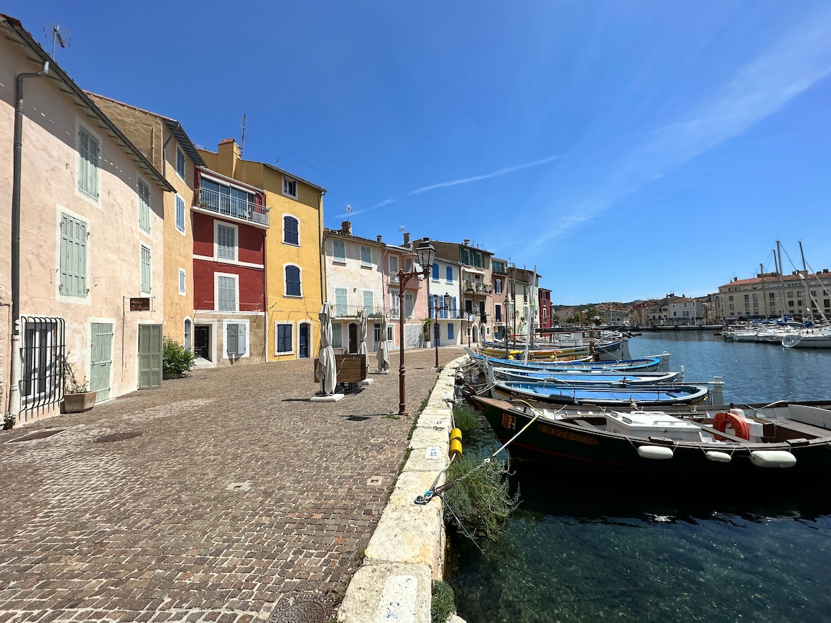 A scenic waterfront view captures colorful buildings lining a cobblestone walkway. Fishing boats are moored along the quay, while clear blue skies reflect over the tranquil waters, highlighting the charm of this vibrant neighborhood.