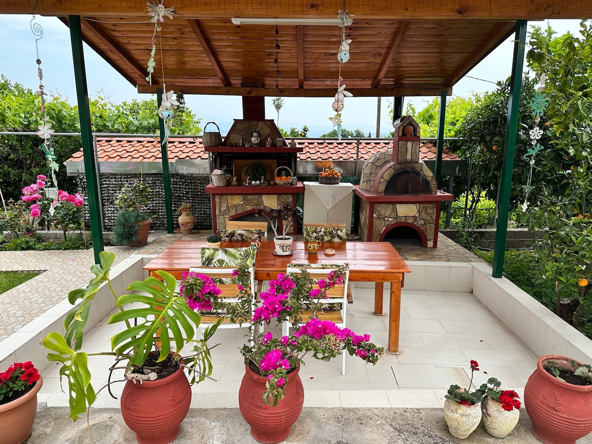 An outdoor dining area is shaded by a wooden roof, featuring a long table arranged with plants and decorative pots. A stone barbeque and a traditional pizza oven stand at the back, surrounded by vibrant flowers and greenery enhancing the relaxing atmosphere.