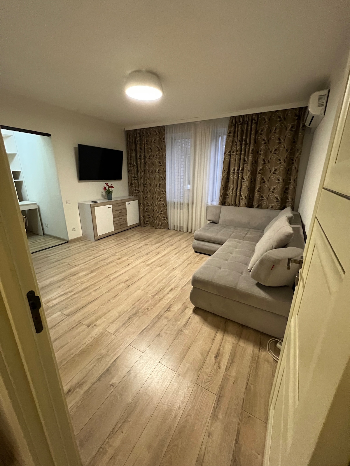 The living area features a light-colored wooden floor and a gray sectional sofa positioned near a wall-mounted television. Natural light enters through large windows, which are adorned with sheer curtains. A small console table is placed against the wall, enhancing the room's functionality.