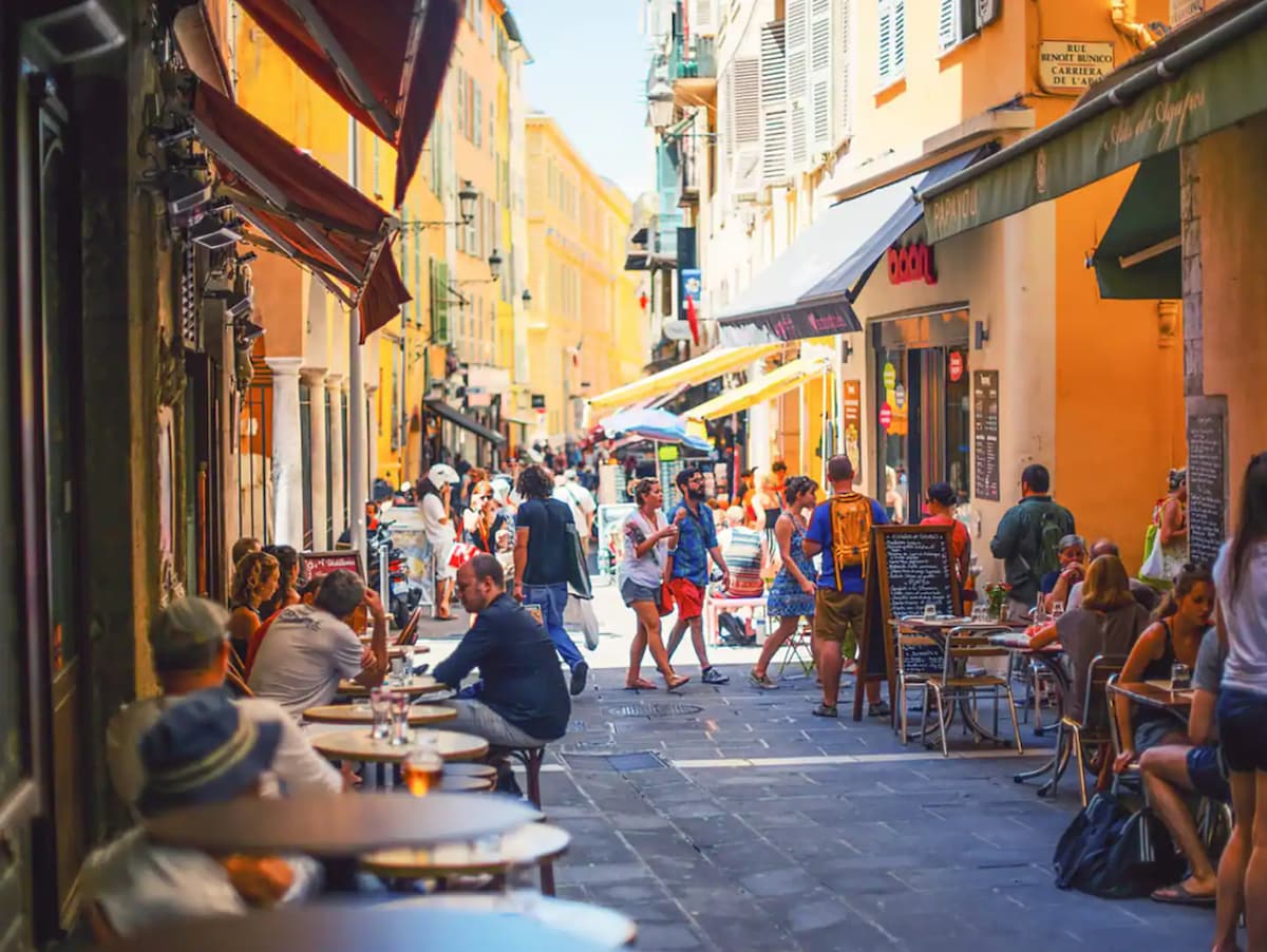 A lively street scene is captured, showing a bustling outdoor dining area with patrons seated at tables. Colorful buildings line the street, and umbrellas provide shade. People can be seen walking between shops and cafes, creating a vibrant atmosphere typical of Nice.