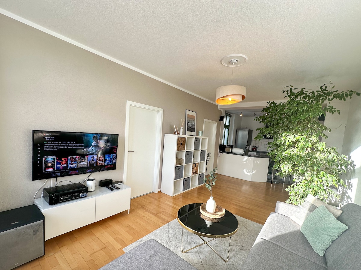 A spacious living area is highlighted by a large TV mounted on the wall. A stylish coffee table is centered on a wooden floor, accompanied by a comfortable sofa. Natural light filters in through a nearby window, complemented by a decorative plant and a modern pendant light above.