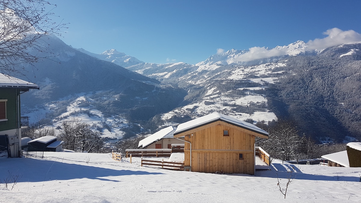 A scenic winter landscape features a wooden chalet surrounded by a blanket of snow. Majestic mountains rise in the background, contrasting with the clear blue sky. The serene setting includes a distant view of neighboring houses, creating a peaceful and inviting atmosphere.