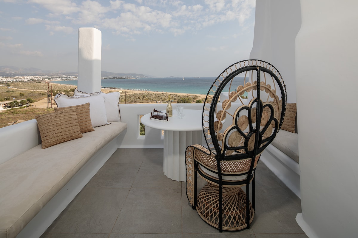 An outdoor terrace features a round table surrounded by a stylish rattan chair and built-in seating. Soft pillows enhance the comfortable seating area, with views overlooking the sea and landscape. The space is defined by smooth, neutral tones and a bright, open atmosphere.