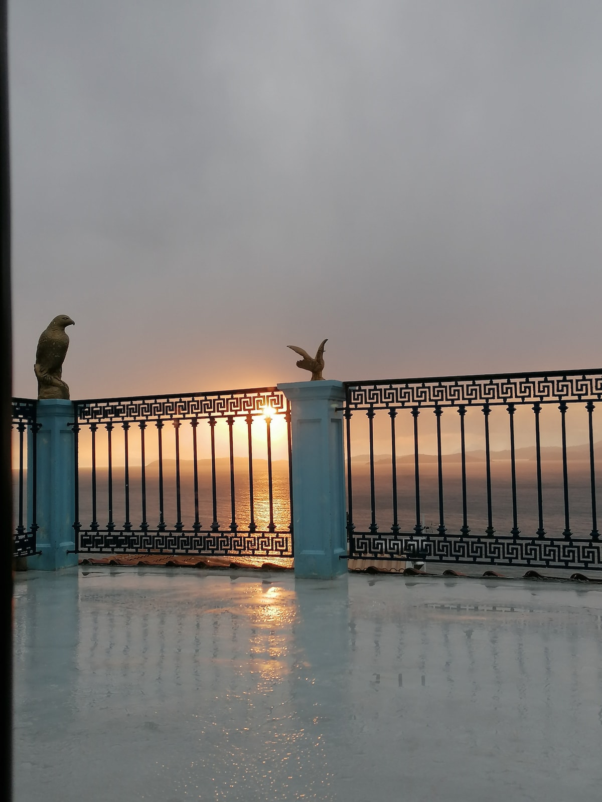 A balcony railing with decorative elements overlooks a serene sea at sunset. The sun is partially hidden behind clouds, casting soft light across the water's surface. The floor is wet, reflecting the light and surrounding scenery, creating a peaceful ambiance.