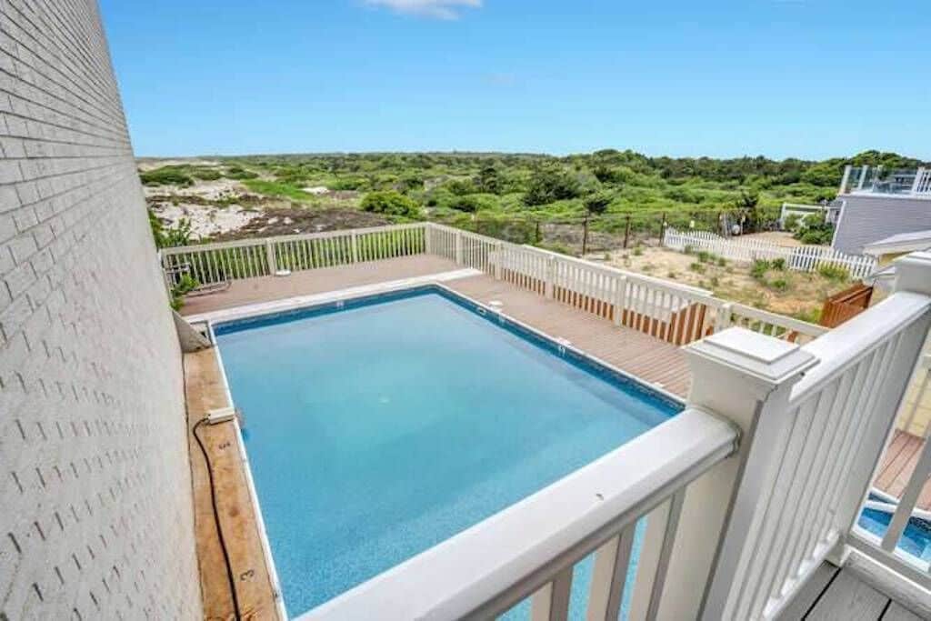 A private outdoor pool is seen surrounded by a wooden deck, offering views of lush greenery and natural landscapes beyond. The clear water of the pool reflects the sky, contributing to a serene, relaxing environment.