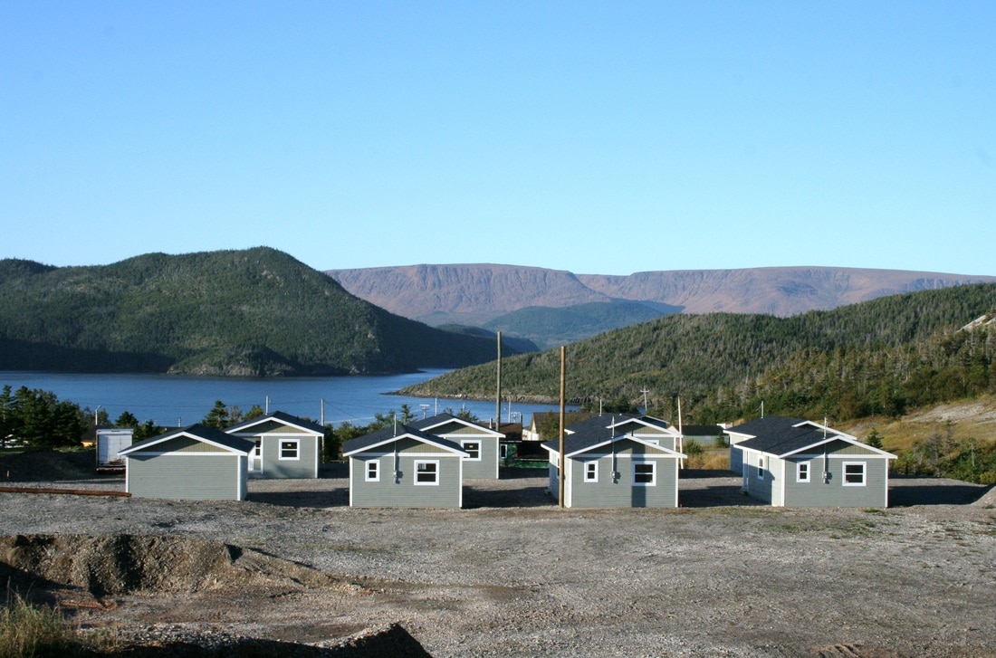 A cluster of light-colored cottages is positioned on a gravel area, with a backdrop of rolling green hills and distant mountains. A calm body of water is visible, reflecting the bright blue sky. The setting conveys a serene and inviting atmosphere.
