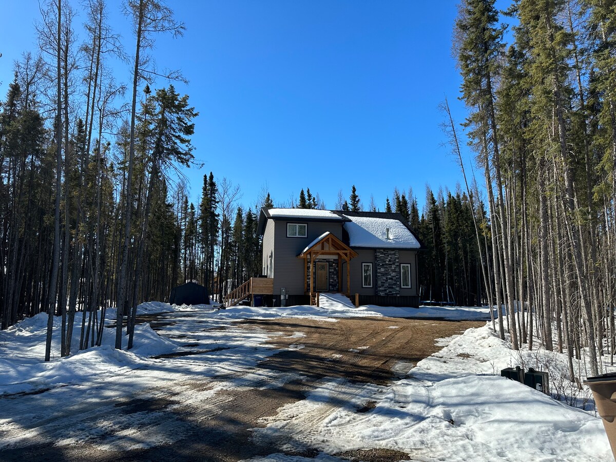 A modern cabin is positioned amidst tall trees, featuring a welcoming entrance with wooden accents. A gravel driveway leads to the cabin, where snow is partially visible on the ground. The clear blue sky contrasts with the greenery surrounding the property.