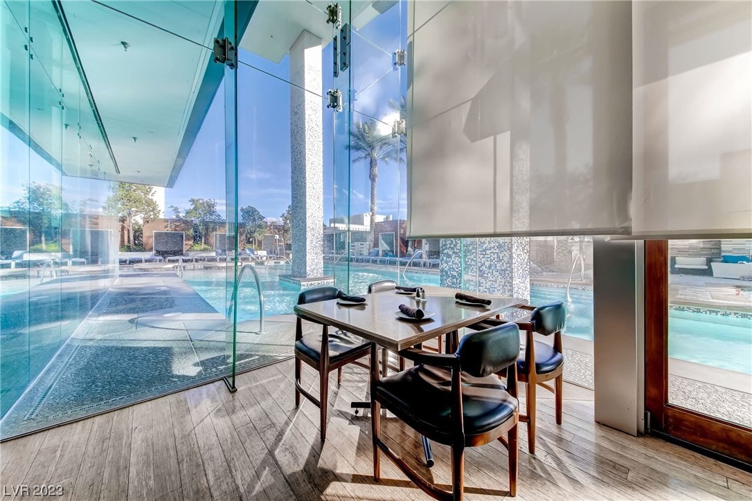 A dining area is positioned behind large glass walls, offering views of the pool and outdoor space. A wooden table is set with four black chairs, while natural light streams through the glass, creating a bright environment.