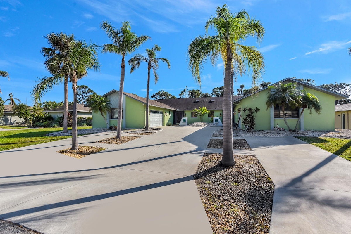 A spacious driveway leads to a light green duplex, flanked by several palm trees. The well-maintained yard features a gravel area and manicured grass, providing a welcoming entrance. An ample parking space is visible, contributing to the serene ambiance of the property.
