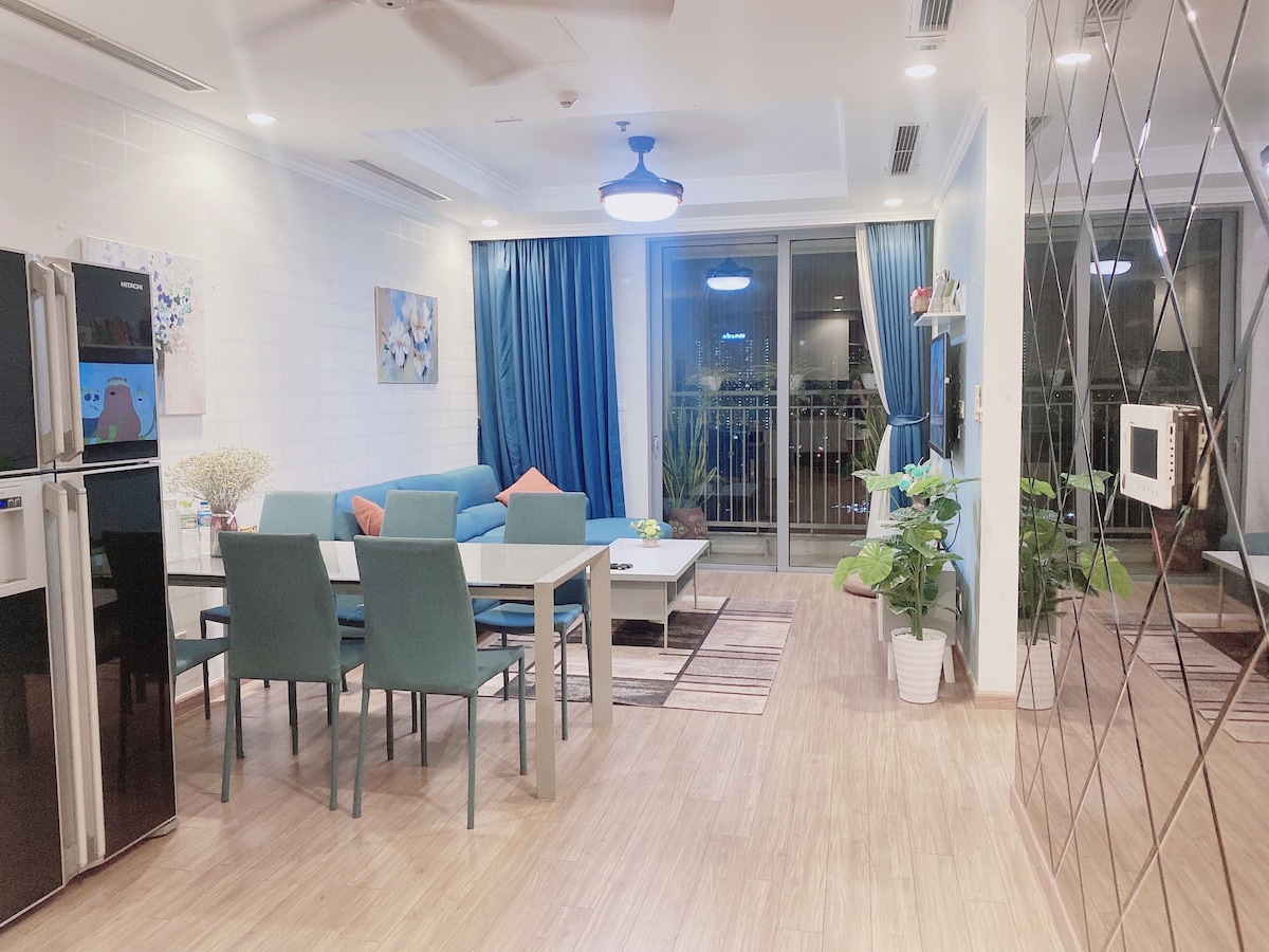 A comfortable living area features a light wood floor and large windows that provide natural light. A modern dining table is surrounded by blue chairs, while a cozy couch with cushions adds to the inviting atmosphere. Potted plants and artwork contribute to the decor.