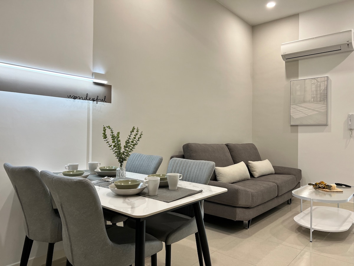 A modern dining area is set with a rectangular table, four chairs, and a decorative centerpiece featuring greenery. A cozy grey sofa is positioned nearby, complemented by neutral-colored cushions. A wall-mounted air conditioning unit and soft lighting enhance the inviting ambiance.
