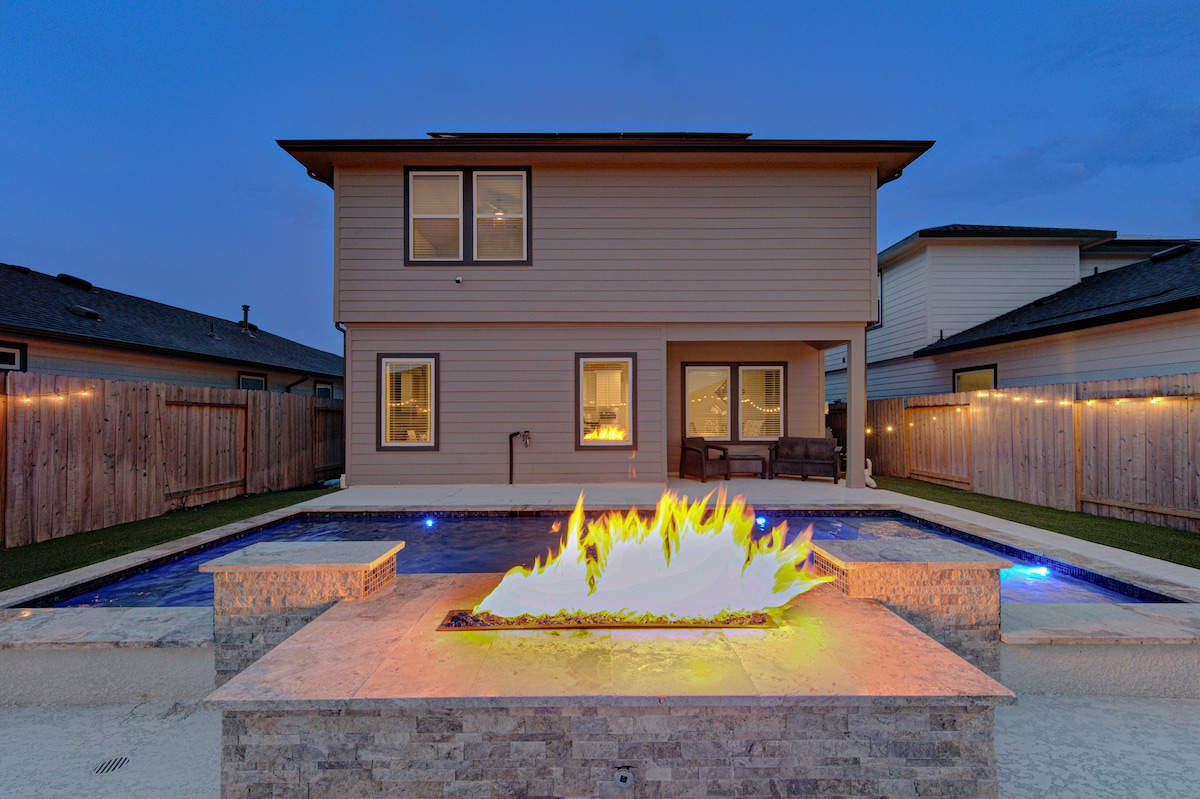 A serene backyard scene is illuminated by a fire feature, surrounded by a stone border. The inviting glow contrasts with the modern two-story home in the background. A tranquil pool reflects soft lighting, enhancing the outdoor space for relaxation and gatherings.