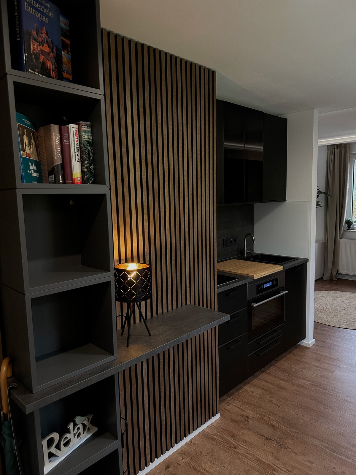 A modern kitchenette features dark cabinetry and sleek appliances, including an oven and a stovetop. A wooden accent wall adds warmth, while a small lamp offers ambient lighting. Open shelving displays books and decorative items, enhancing the functional yet stylish space.