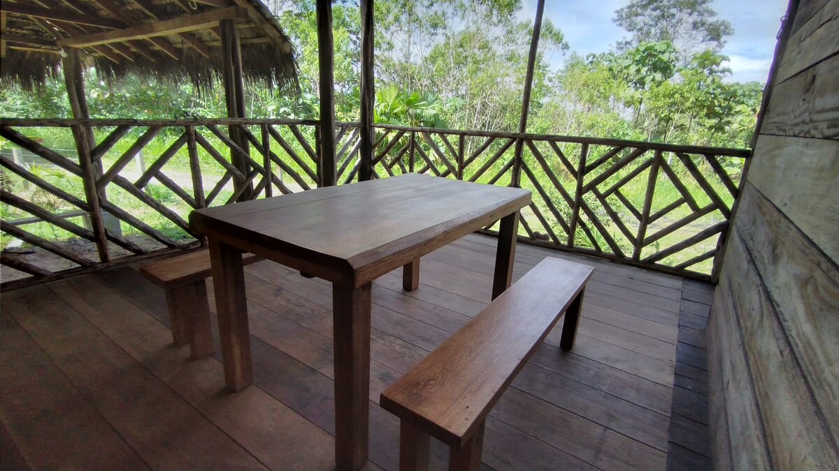 A wooden dining table is positioned within a screened-in area, accompanied by two matching benches. The space features open walls that allow for views of the surrounding greenery and the natural light to enter comfortably.