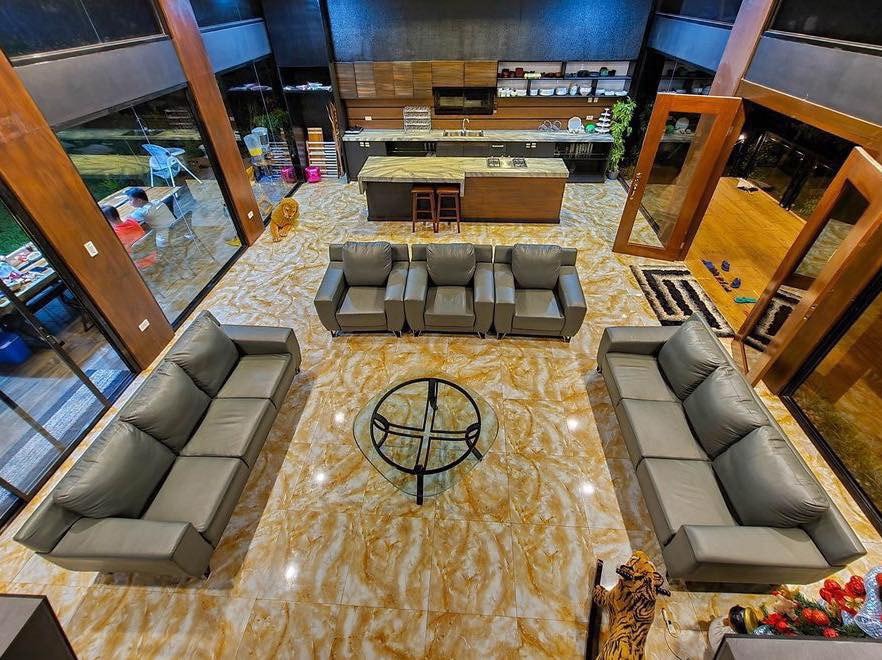A spacious living area is presented with two large sofas and two armchairs arranged around a circular glass coffee table. Large glass walls allow natural light to fill the space, while a modern kitchen with wooden accents is visible in the background.