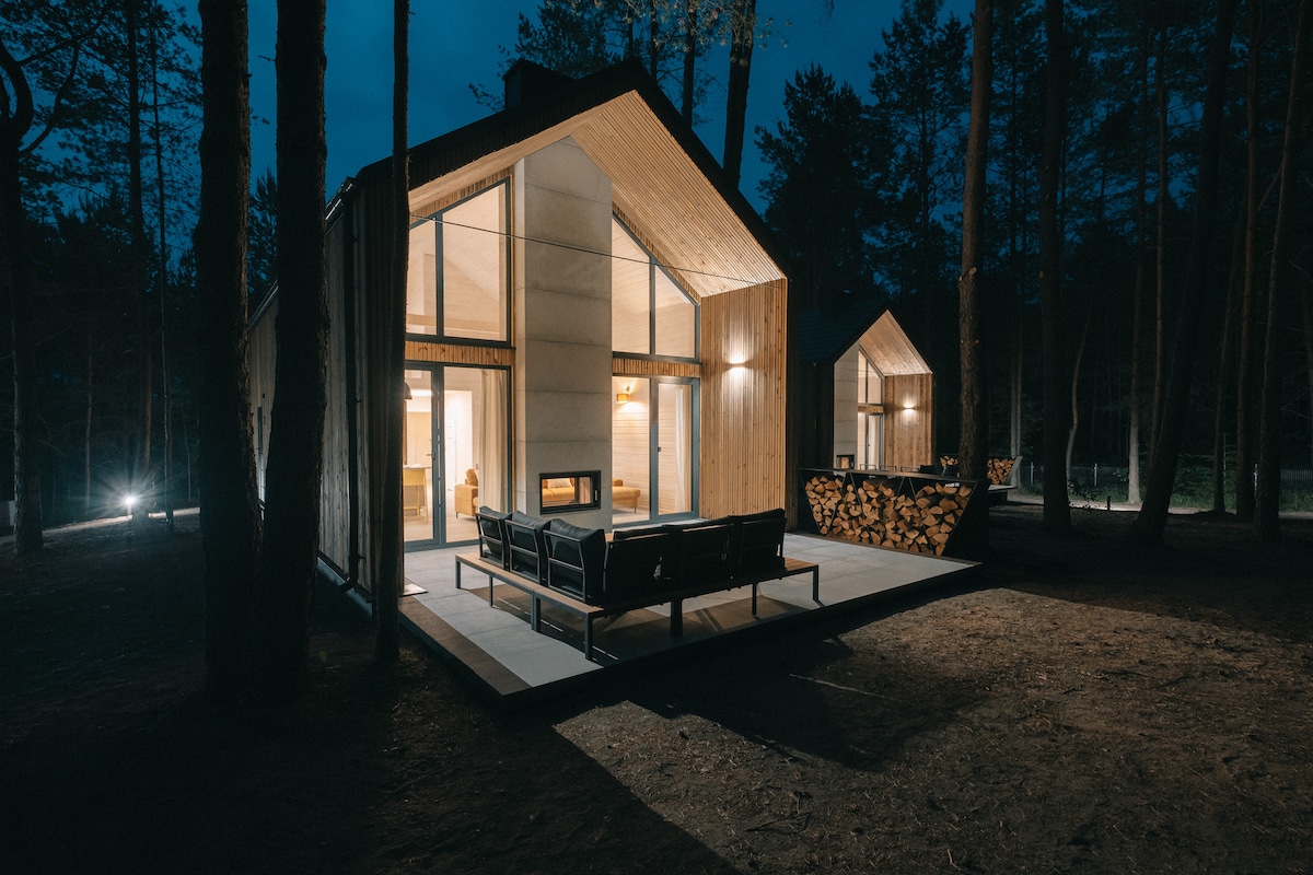 The exterior of the Amber house is seen at night, featuring a modern design with large glass windows. A welcoming terrace area includes seating and a woodpile. The surrounding forest is softly illuminated, creating a serene atmosphere.