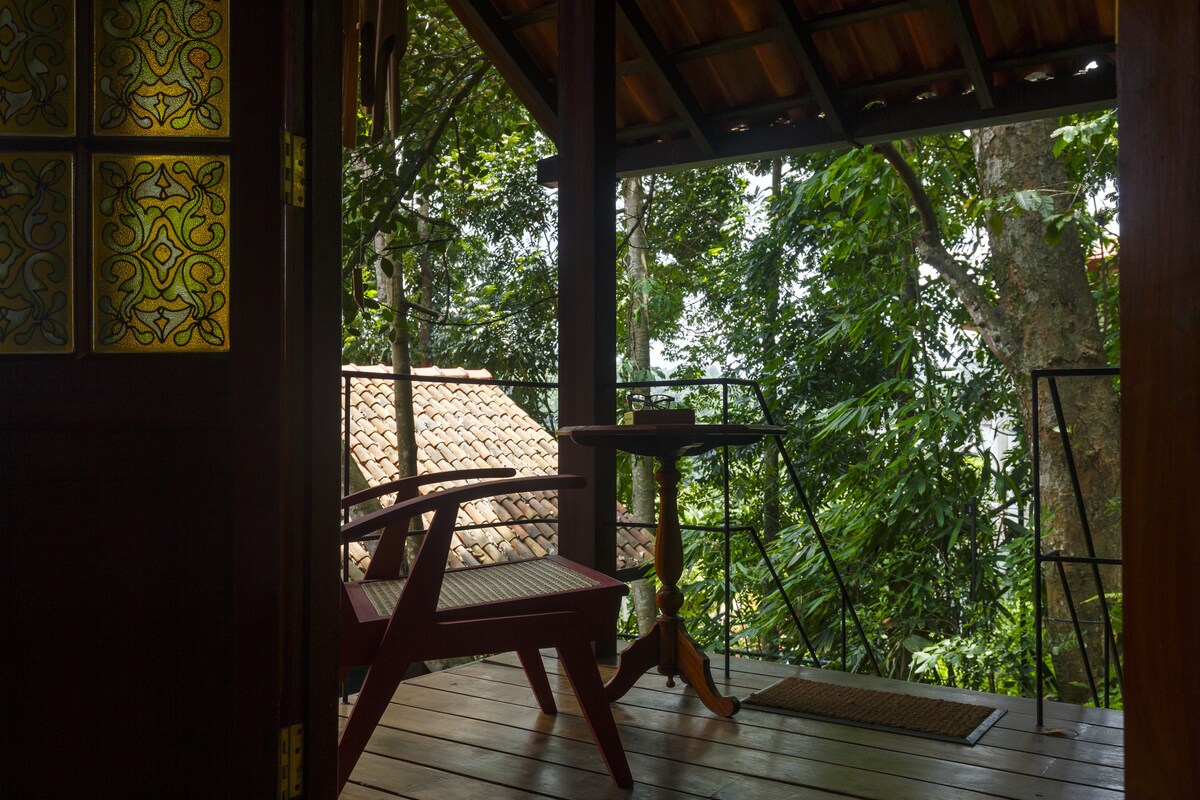 A covered balcony with wooden flooring features a red chair and small round table. Green foliage surrounds the area, providing a natural backdrop. Decorative glass panels on the door add a touch of artistry while allowing light to filter through.