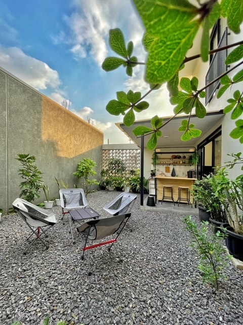 An outdoor sitting area is surrounded by lush greenery, featuring several chairs positioned on gravel. A bar-like counter is visible in the background, adorned with decorative plants and shelves. Soft natural light filters through the plants, creating a serene atmosphere.