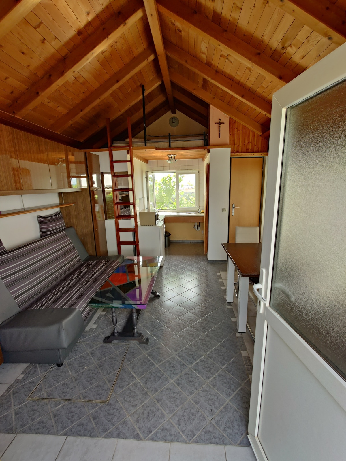 An open living area features a high wooden ceiling with exposed beams. A glass table is positioned beside a gray sofa bed, while a ladder leads to a loft space. A window provides natural light and views of the surroundings.