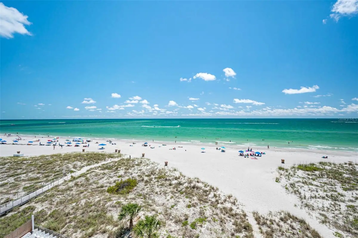 A wide view of Madeira Beach showcases soft white sands meeting the turquoise waters of the Gulf. Sunbathers lounge on colorful beach chairs under a clear blue sky, while gentle waves lap at the shore's edge. Dunes with greenery are visible in the foreground.