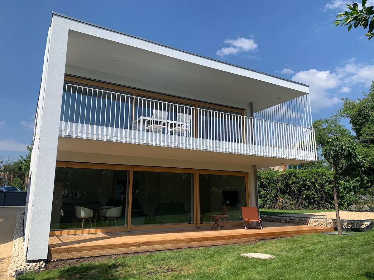 A modern, two-story house features a large balcony with white outdoor furniture and glass railings. The ground level features expansive glass doors opening onto a wooden deck, surrounded by a well-maintained garden with greenery and a small tree.