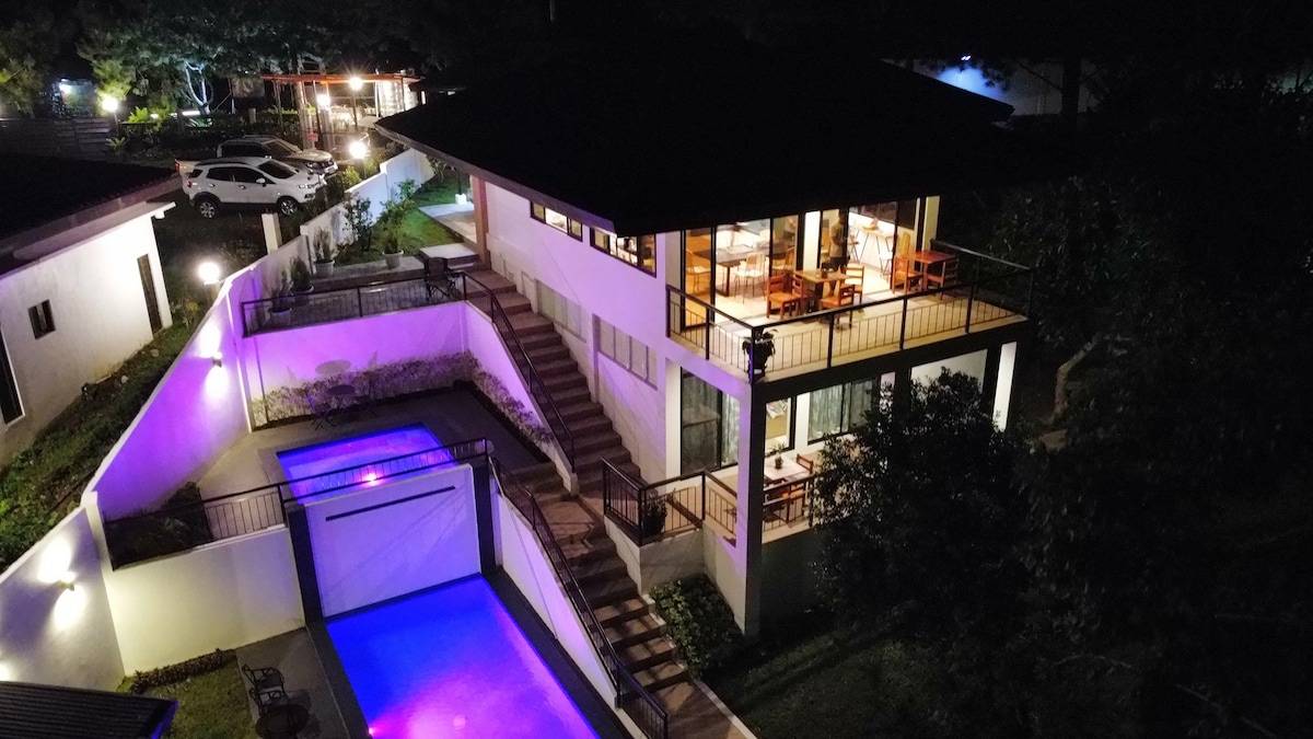 The two-story home is positioned on a hillside, featuring a spacious outdoor area with a swimming pool illuminated in blue light. Multiple pathways lead to the entrance, surrounded by lush greenery and softly glowing outdoor lights.