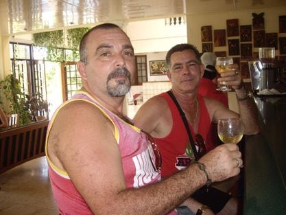 Two individuals are seated at a bar, each holding a cocktail glass. The bar features a polished surface with wooden accents and a collection of framed artwork in the background. Natural light filters through large windows, contributing to a relaxed atmosphere.