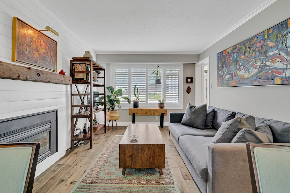 The spacious living area features a comfortable gray sofa arranged around a rustic wooden coffee table. A wall-mounted artwork and a wooden bookshelf accentuate the space, while large windows allow natural light to fill the room. Houseplants add a touch of greenery.