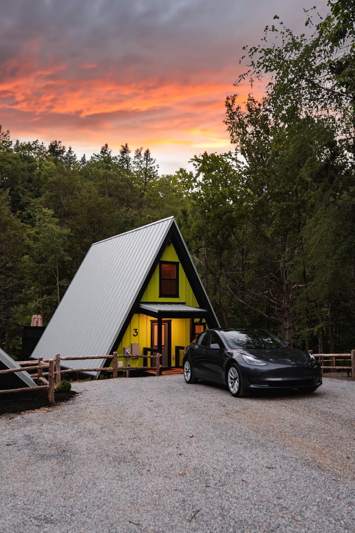An eco-friendly A-Frame retreat is depicted amidst lush greenery, featuring a striking exterior with a modern design. A black vehicle is parked on the gravel driveway, showcasing convenient access. The evening sky is illuminated with hues of orange and pink, creating a serene backdrop.