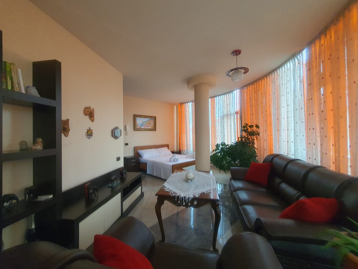 A well-lit living area showcases large windows draped with sheer orange curtains. A comfortable seating arrangement is complemented by a decorative table and a potted plant. A spacious bed is positioned in the background, surrounded by light-colored walls and tasteful decor.