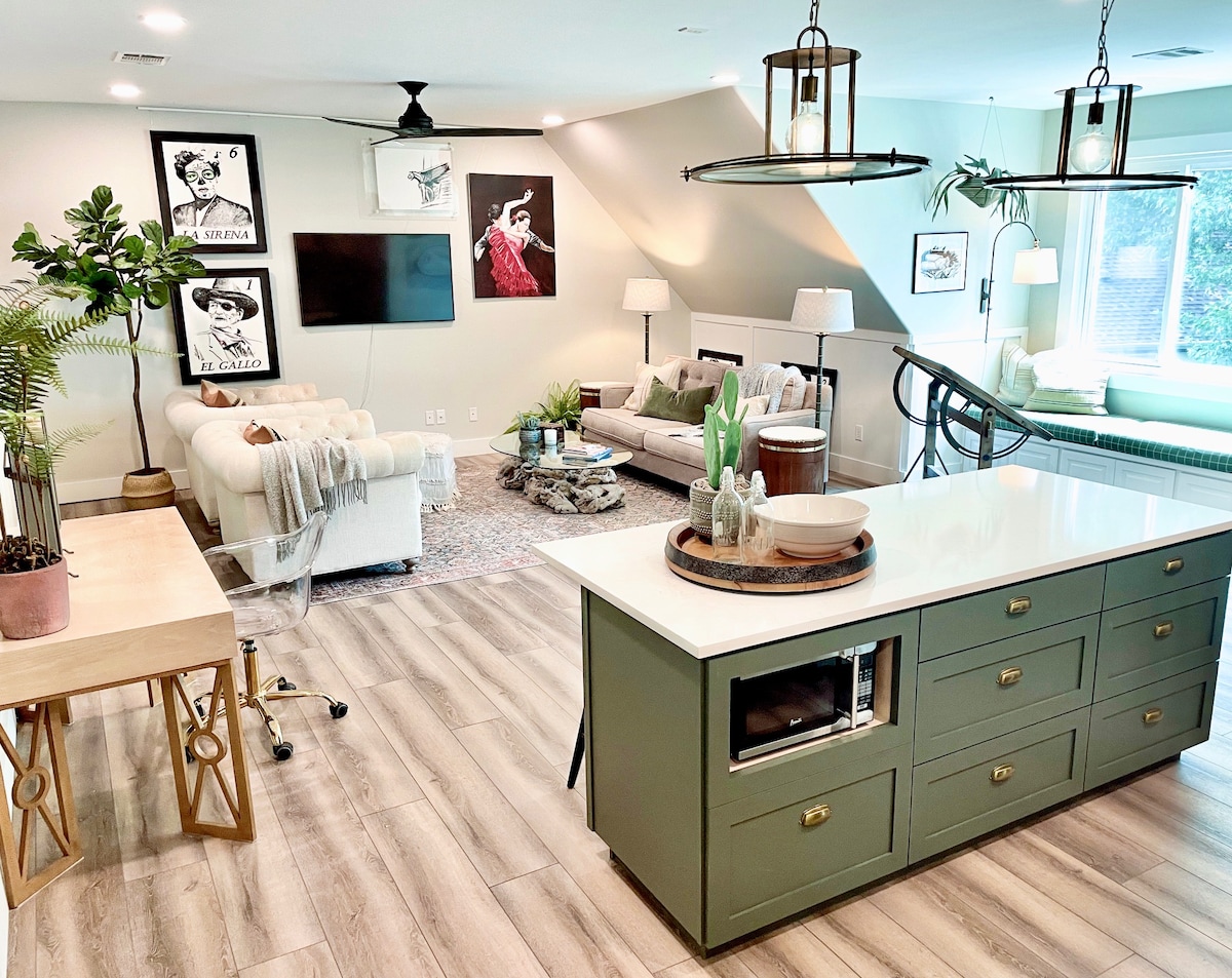 A stylish living area features a light-colored sofa, a decorative coffee table, and wall art adorned with vintage-inspired prints. Natural light illuminates the room, and a dining table is positioned nearby. A modern kitchen island showcases cabinetry and a microwave, enhancing functionality.