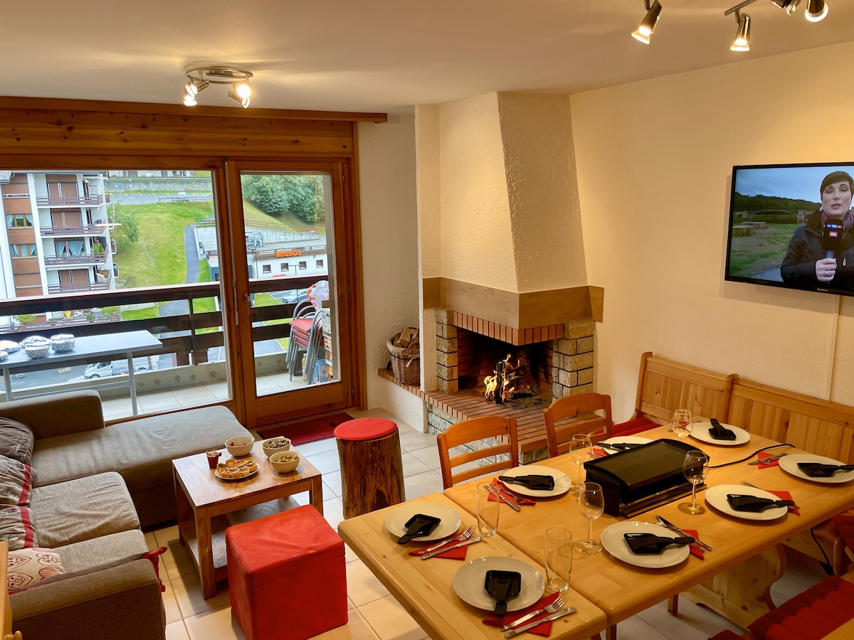 A spacious living and dining area features large windows opening to a balcony, allowing plenty of natural light. A modern fireplace is positioned between a comfortable sofa and a wooden dining table set for six, with additional seating offered by red ottomans.