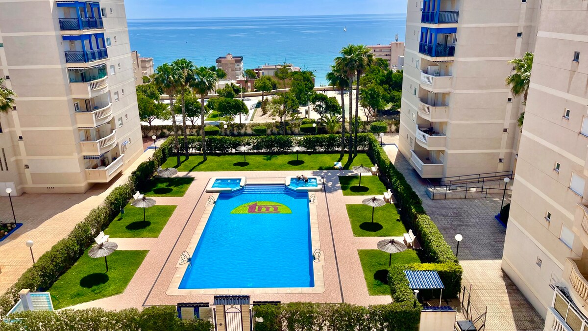 This aerial view showcases the communal swimming pool surrounded by neatly manicured lawns and palm trees. The pool is set against a backdrop of the Mediterranean Sea, offering a tranquil atmosphere and scenic views. Loungers and umbrellas provide space for relaxation around the pool area.