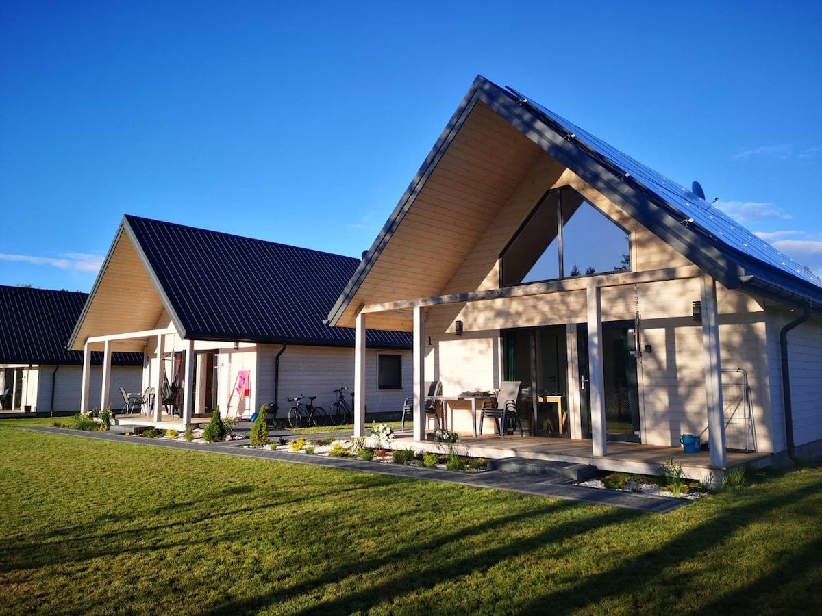 Two modern cabins are shown with sloped roofs and large front porches. The outdoor area features a well-kept lawn and decorative landscaping. Bicycles are parked at the side, suggesting opportunities for outdoor activities. Clear blue skies complement the scene.