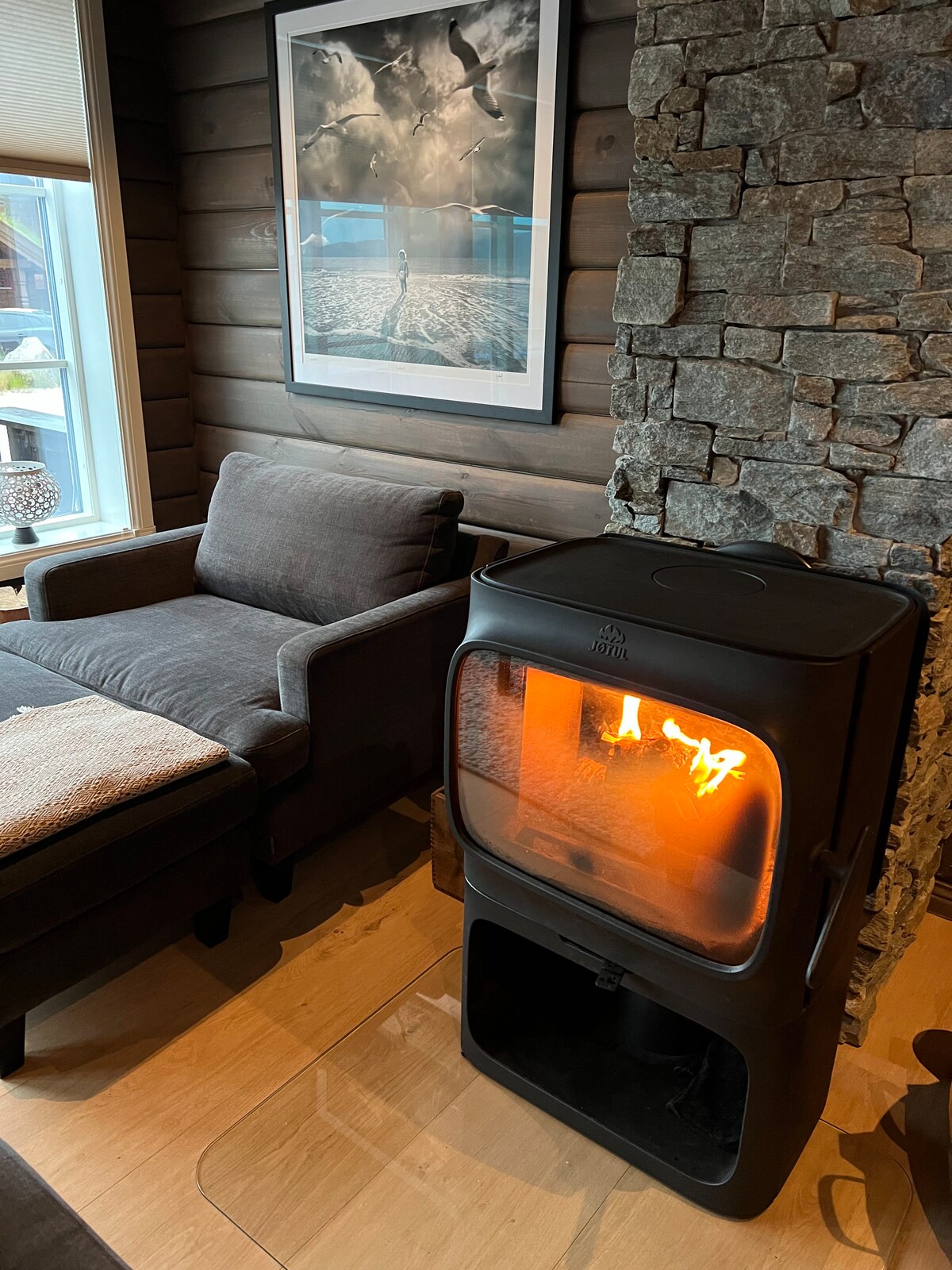 A modern wood stove with a glass front displays a warm flame, set against a backdrop of natural stone walls. A cozy seating area, including a gray sofa, complements the inviting ambiance. Large windows allow light to filter into the space.