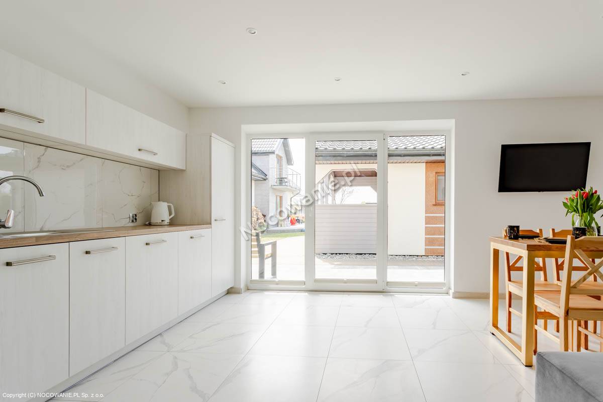 A modern kitchen area features sleek cabinetry with a smooth countertop and a sink. Large glass doors provide natural light and views of the outdoor space. A small dining table with wooden chairs is visible, enhancing the functionality of the room.