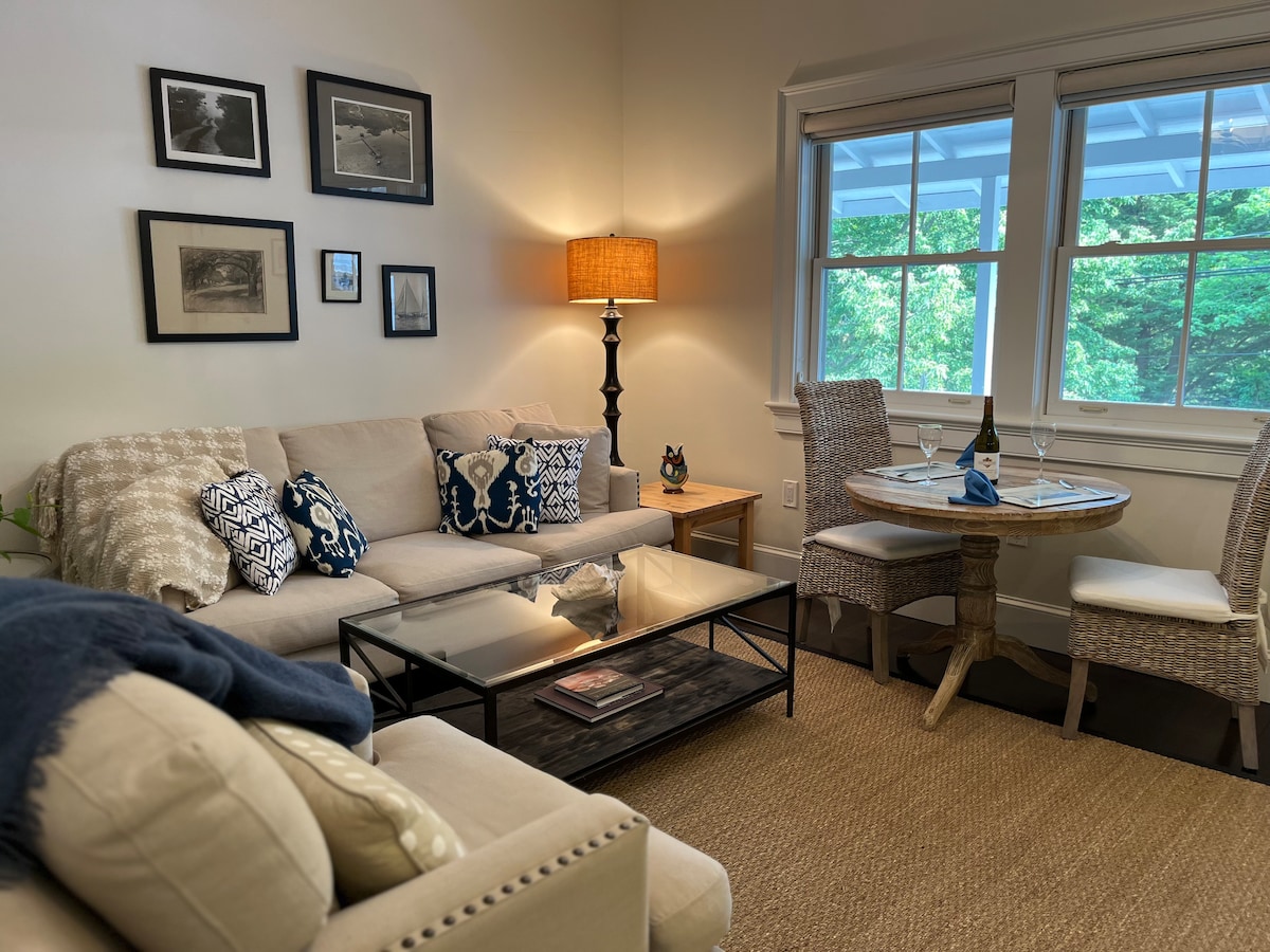 A comfortable living area is furnished with a light-colored sofa and accent pillows, complemented by a cozy armchair. A stylish lamp illuminates the space, while a glass-top coffee table is paired with a modest dining table for two, surrounded by woven chairs. Natural light enters through two large windows.