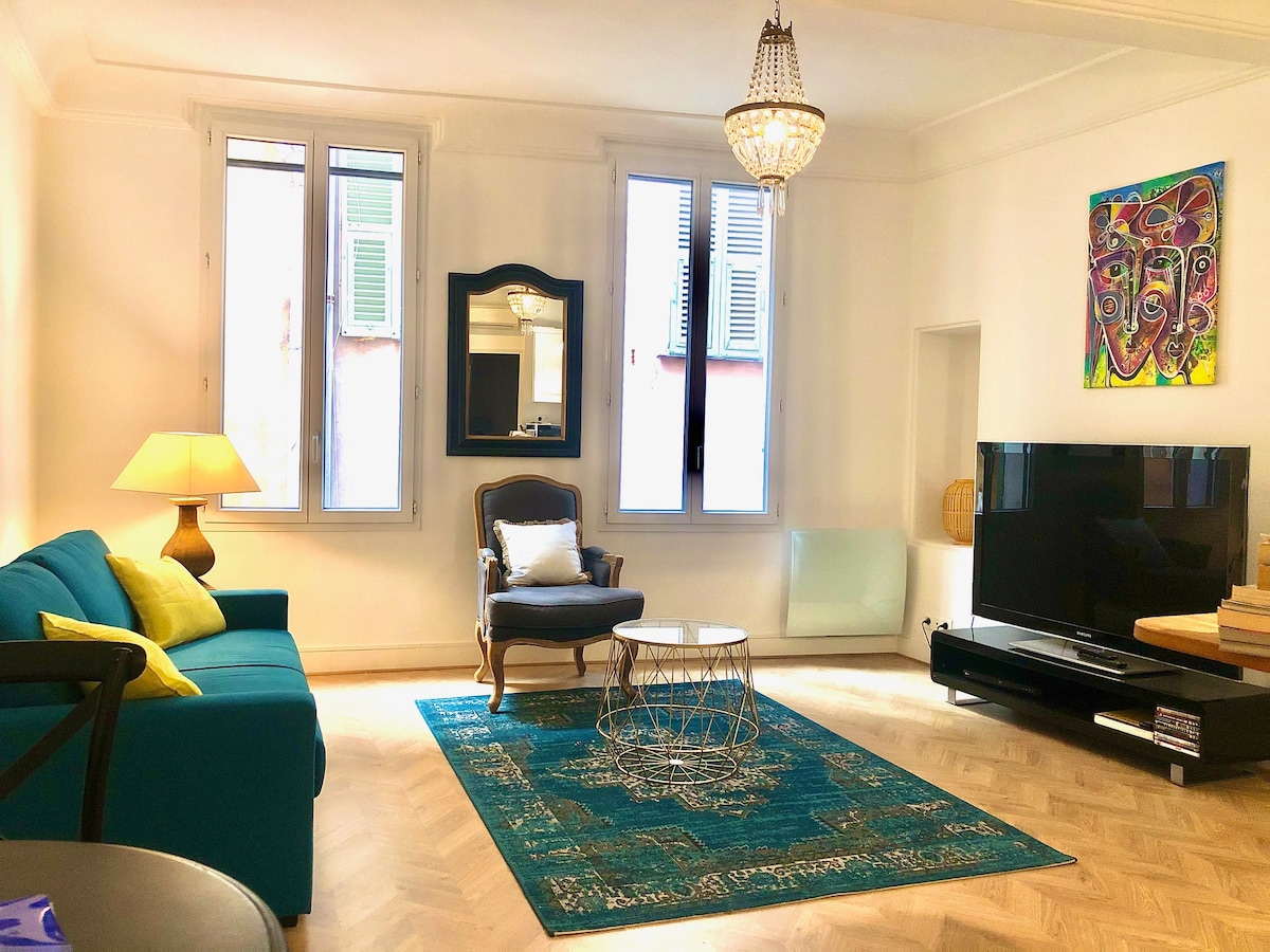 A bright living area features a comfortable couch adorned with vibrant cushions and a stylish armchair. Large windows allow natural light to fill the space, complemented by a decorative mirror and a colorful artwork. A modern television is positioned on a sleek media console.