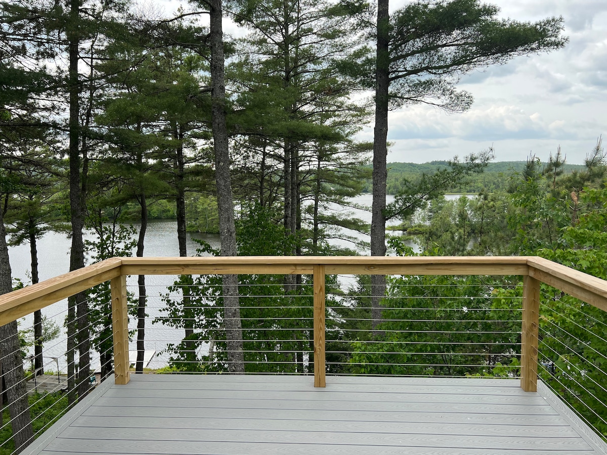A wooden deck is presented, surrounded by tall trees and offering views of the serene lake below. The deck's railing features metal wiring, providing a modern aesthetic while maintaining unobstructed sightlines to the lush surroundings.