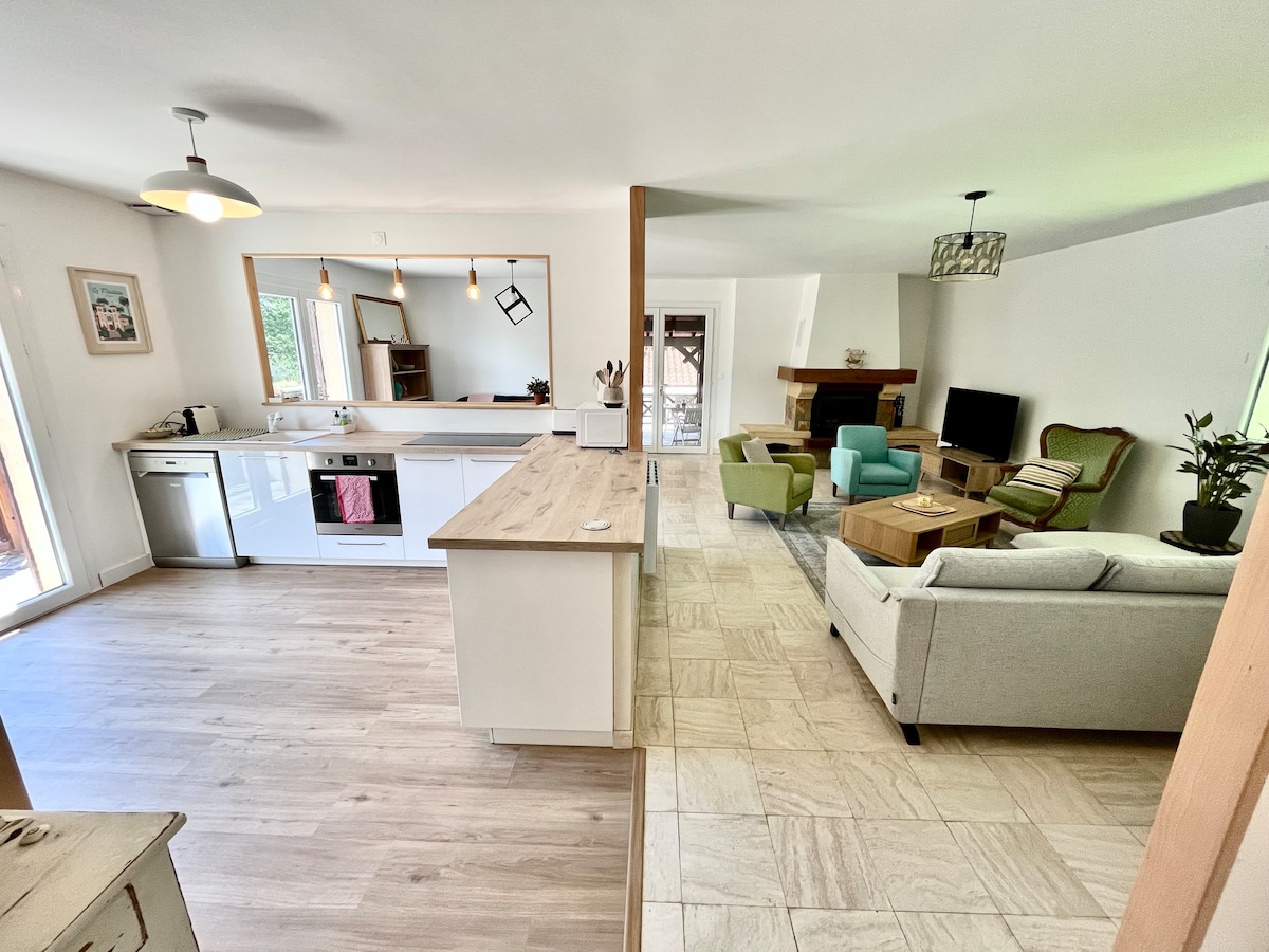 An open-plan living space features a modern kitchen and a comfortable seating area. Natural light floods in, highlighting a soft-colored sofa and stylish armchairs arranged around a wooden coffee table. A television sits in front of a fireplace, adding to the welcoming ambiance.