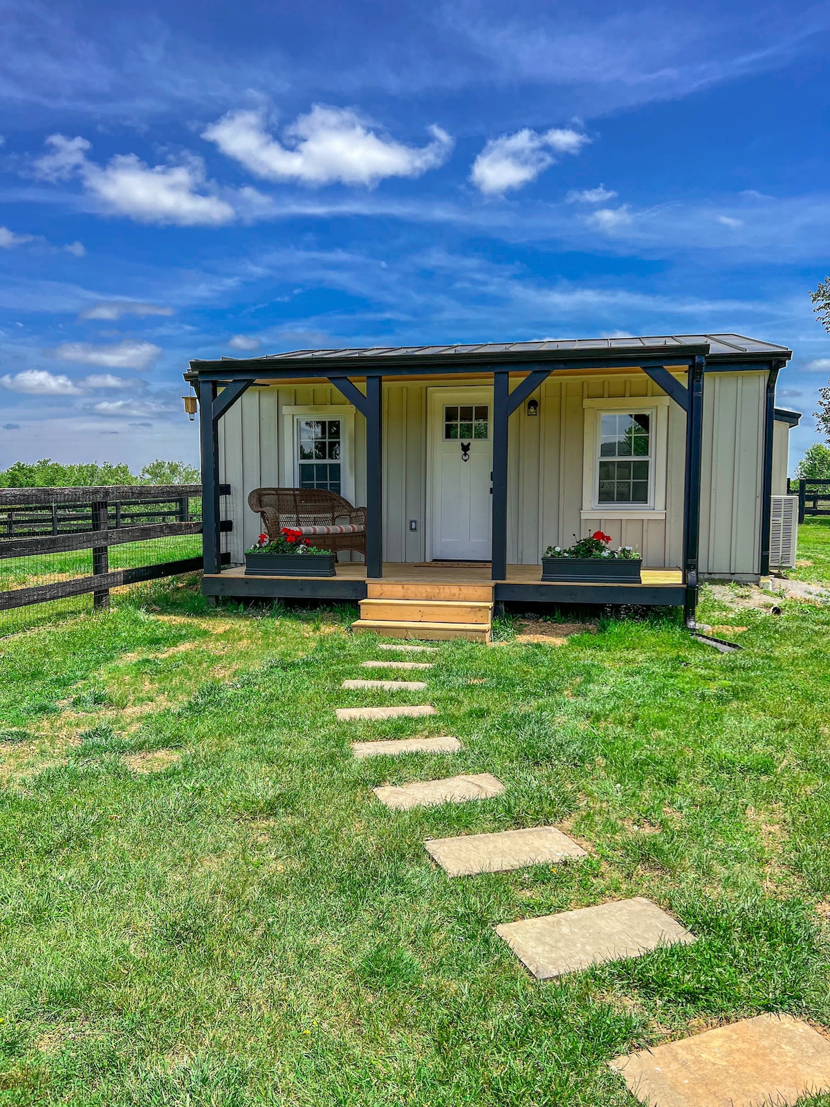 The exterior of a cozy bungalow is presented, with a welcoming porch featuring a wooden swing and flower planters. A stone pathway leads to the door, surrounded by well-maintained grass and a fenced area, all set against a clear blue sky.