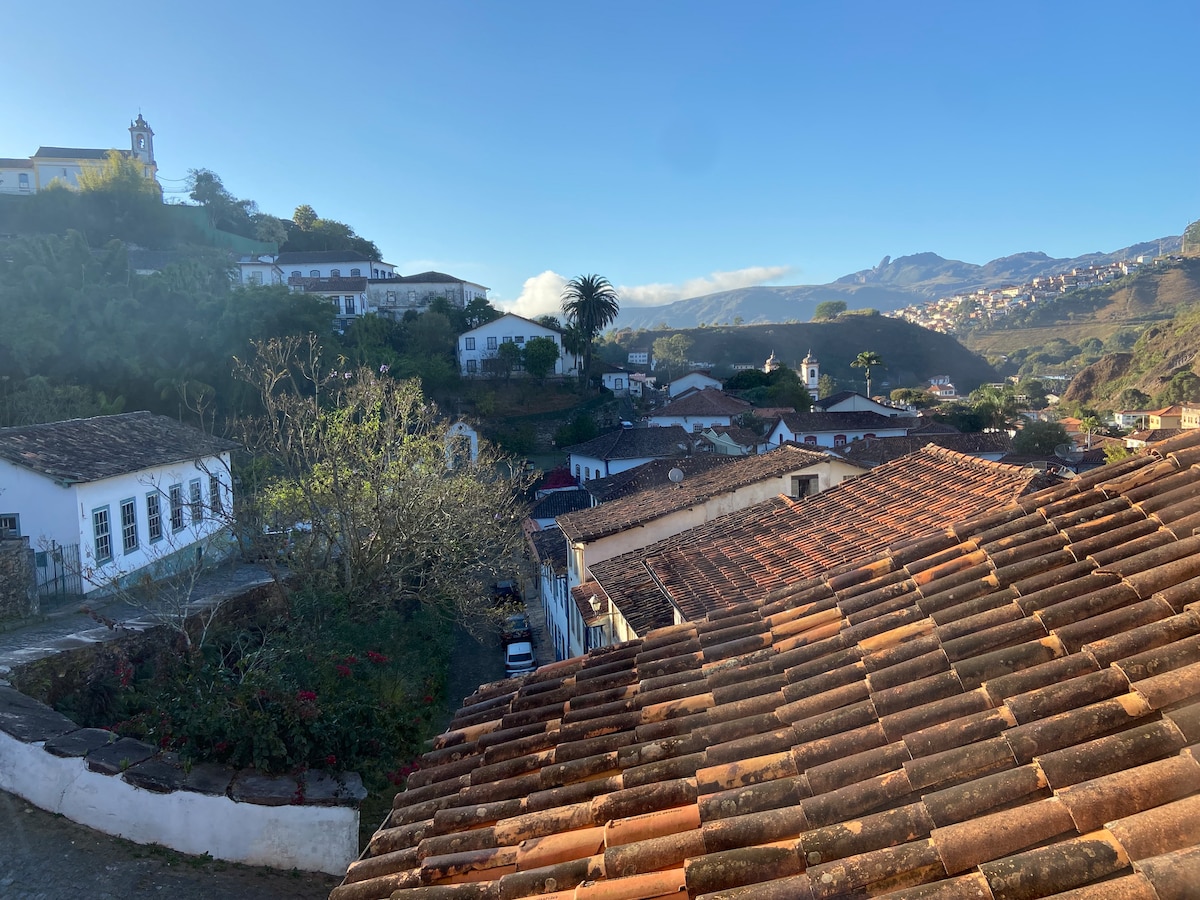 A view of Ouro Preto's hilly landscape is showcased, with colonial-style houses and tiled roofs in the foreground. Lush greenery and distant mountains fill the background, while a clear sky above enhances the scenic beauty of the area.